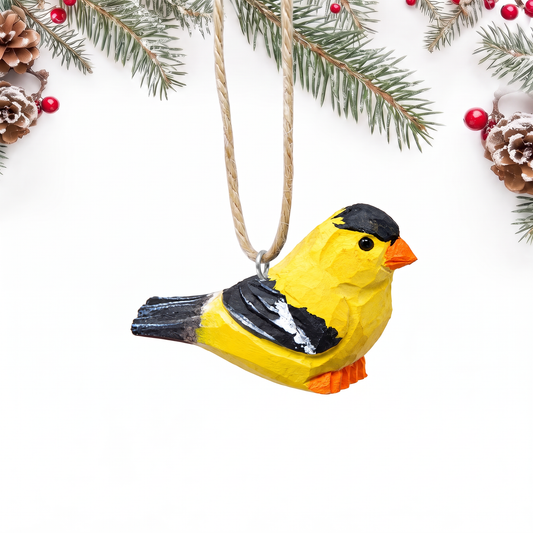 Hand-carved wooden American Goldfinch ornament with a bright yellow body, black cap, black and white wings and an orange beak and feet, hanging by twine from a silver eye hook in the middle of its back, on a white background with pine branches.