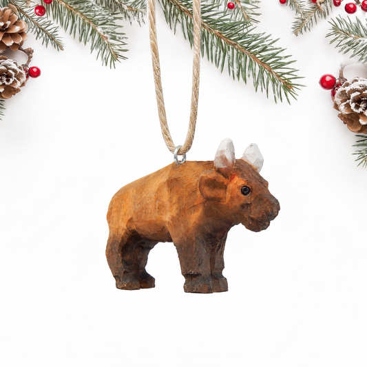 Hand-carved wooden Bison ornament standing, with a brown shaggy body, dark legs and small white horns, hanging by twine from a silver eye hook on its back, on a white background with pine branches.