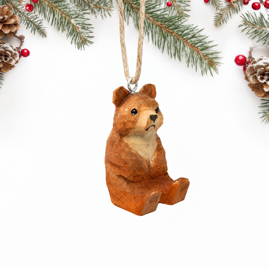 Hand-carved wooden Brown Bear ornament seated, with a brown carved coat and a cream chest and muzzle, hanging by twine from a silver eye hook on top of its head, on a white background with pine branches.