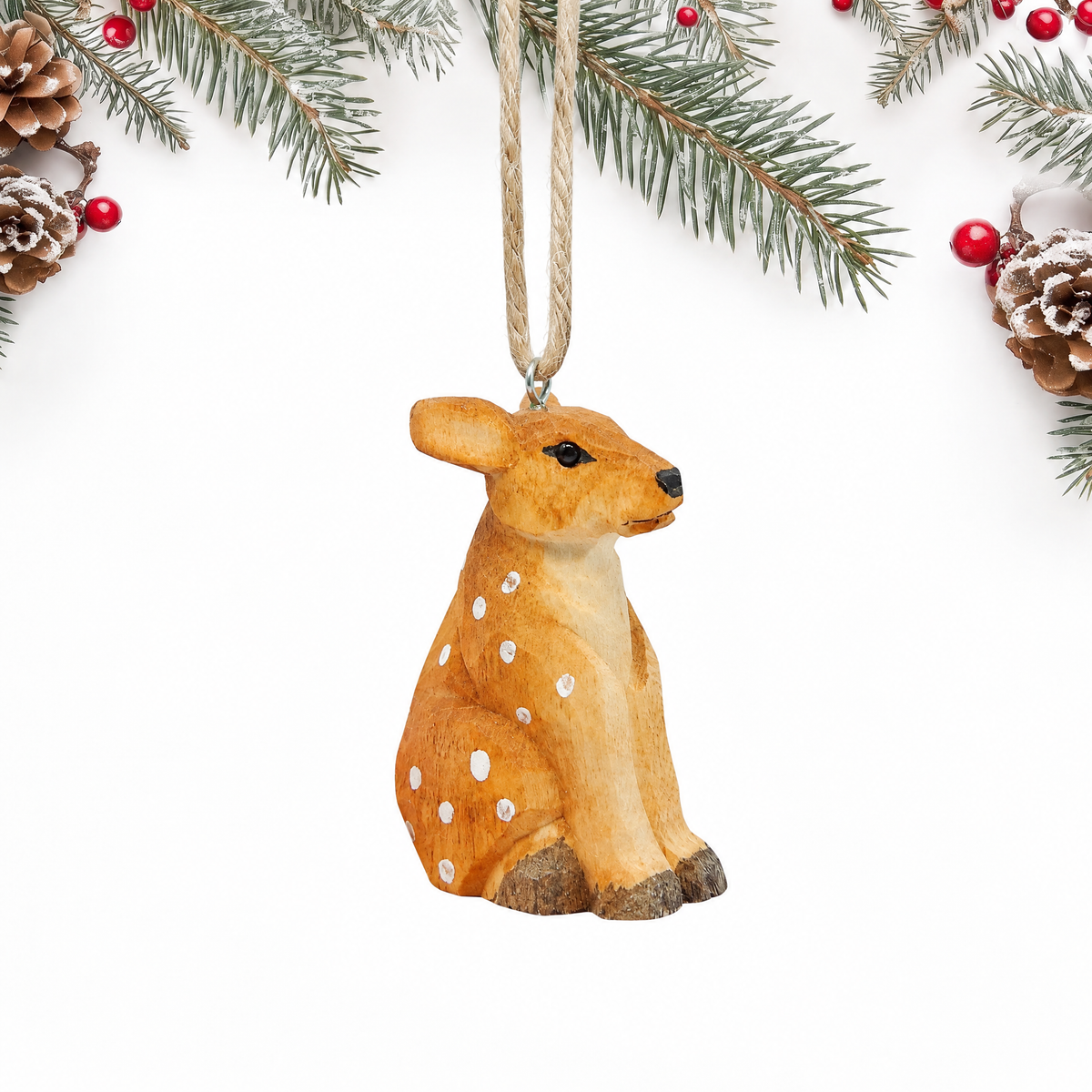 Hand-carved wooden Deer Fawn ornament seated, with an orange tan coat, white spots, large ears and dark hooves, hanging by twine from a silver eye hook on top of its head between the ears, on a white background with pine branches.