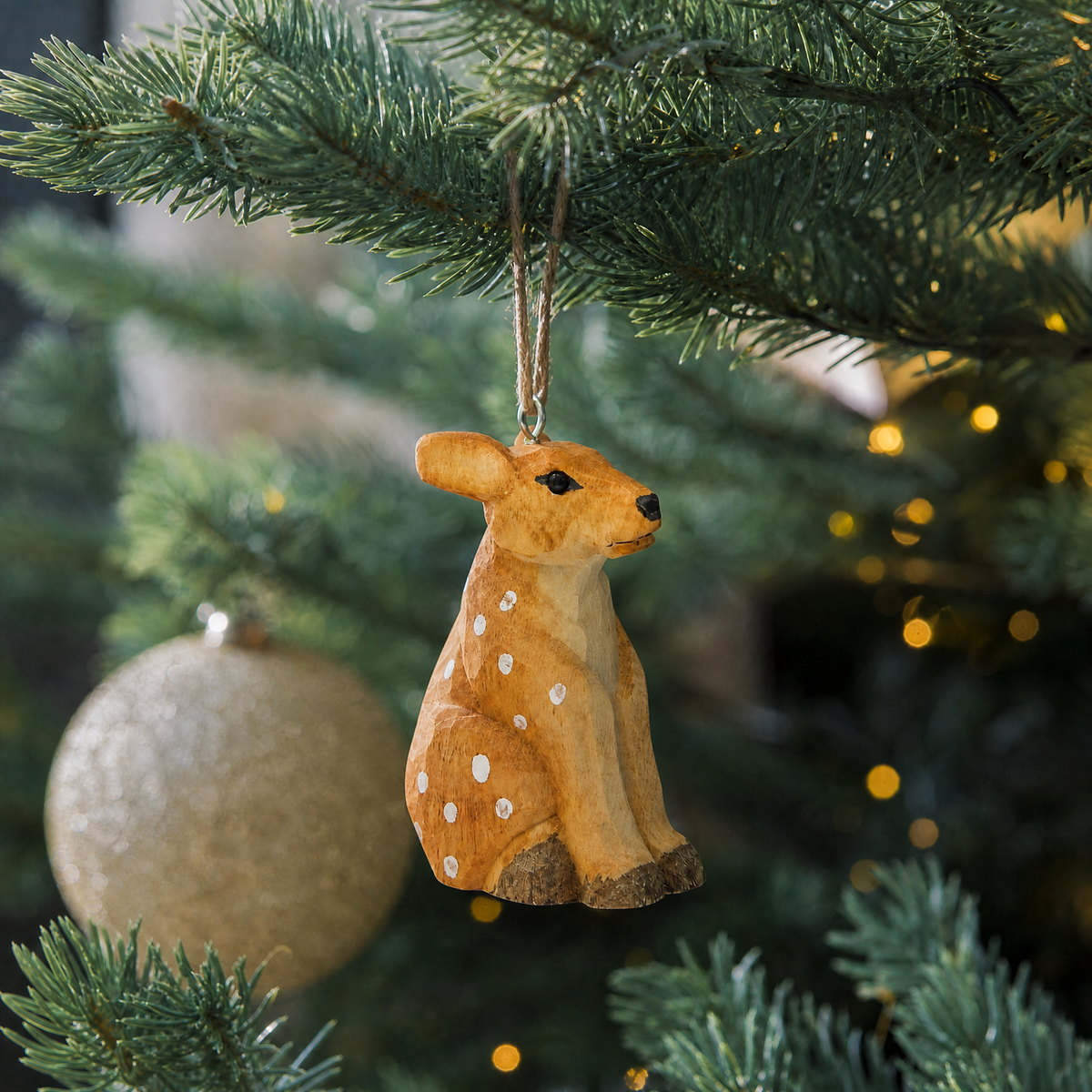 Deer Fawn wooden ornament hanging by natural twine from a Christmas tree branch, with warm string lights and a gold glitter ball ornament behind it.