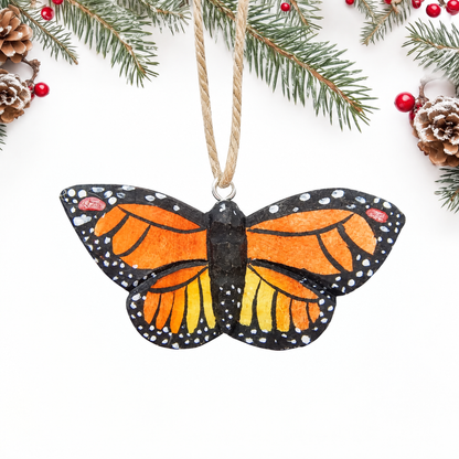 Hand-carved wooden Monarch Butterfly ornament with open orange wings, black veins and borders, white dots and a black body, hanging by twine from a silver eye hook at the top of its body, on a white background with pine branches.