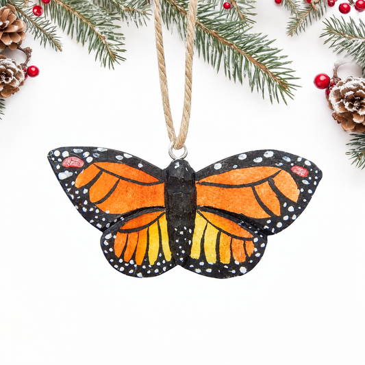 Hand-carved wooden Monarch Butterfly ornament with open orange wings, black veins and borders, white dots and a black body, hanging by twine from a silver eye hook at the top of its body, on a white background with pine branches.