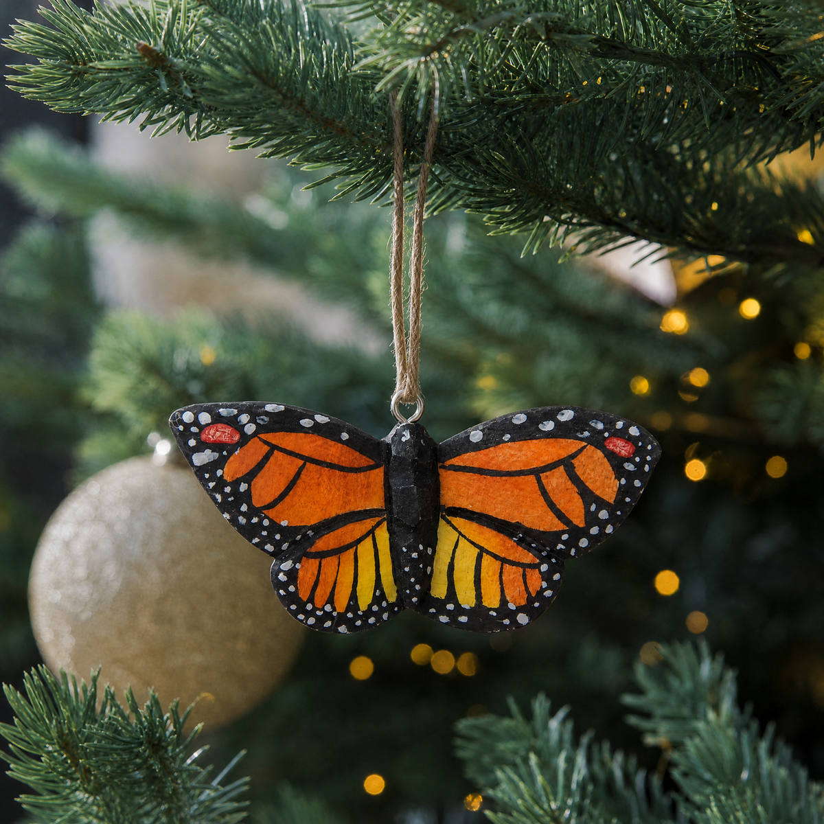 Monarch Butterfly wooden ornament hanging by natural twine from a Christmas tree branch, with warm string lights and a gold glitter ball ornament behind it.