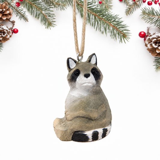 Hand-carved wooden Raccoon ornament seated, with a gray body, black eye mask, white face and a black and white striped tail, hanging by twine from a silver eye hook on top of its head, on a white background with pine branches.