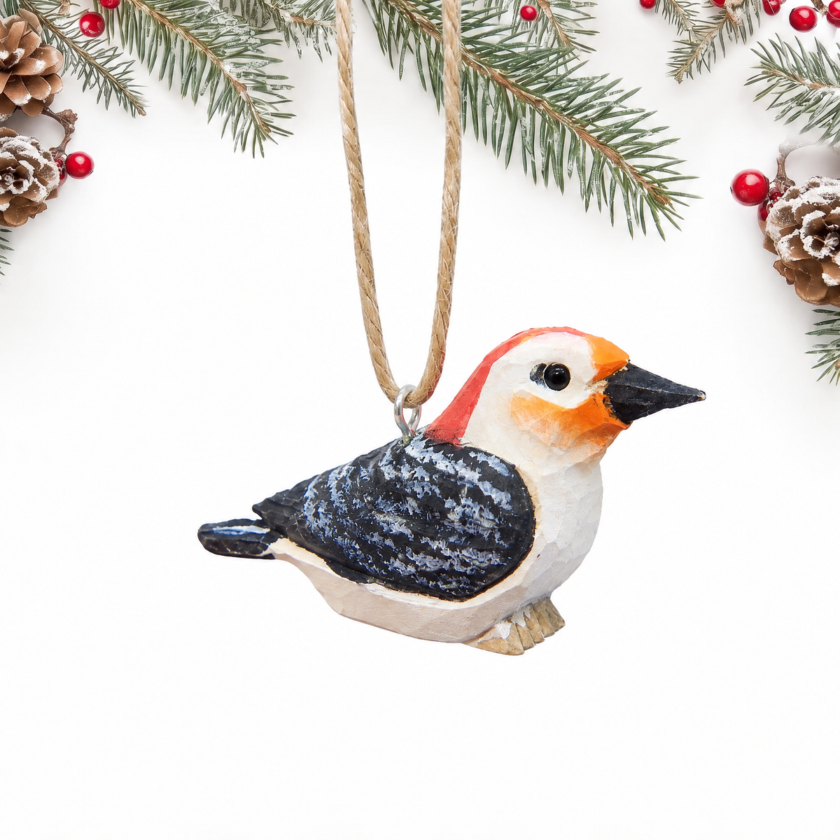 Hand-carved wooden Red-Bellied Woodpecker ornament with a red cap, white face, orange cheek and a black and white barred back, hanging by twine from a silver eye hook in the middle of its back, on a white background with pine branches.