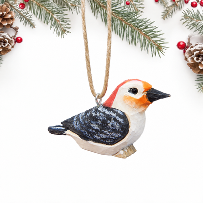 Hand-carved wooden Red-Bellied Woodpecker ornament with a red cap, white face, orange cheek and a black and white barred back, hanging by twine from a silver eye hook in the middle of its back, on a white background with pine branches.