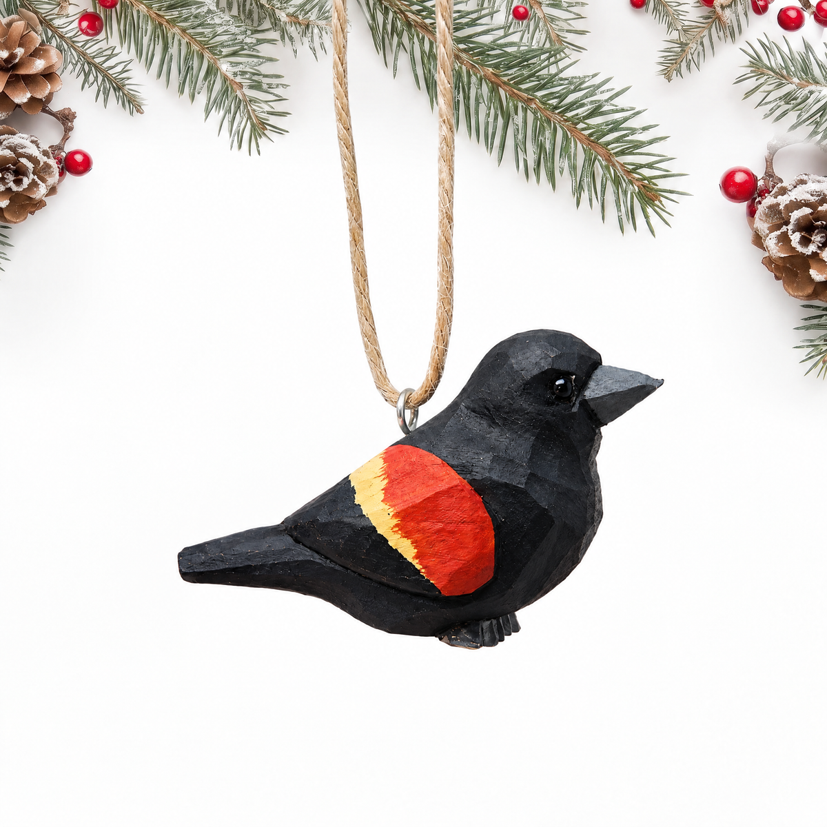 Hand-carved wooden Red-Winged Blackbird ornament with a glossy black body and a red and yellow wing patch, hanging by twine from a silver eye hook in the middle of its back, on a white background with pine branches.