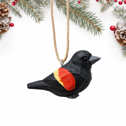 Hand-carved wooden Red-Winged Blackbird ornament with a glossy black body and a red and yellow wing patch, hanging by twine from a silver eye hook in the middle of its back, on a white background with pine branches.