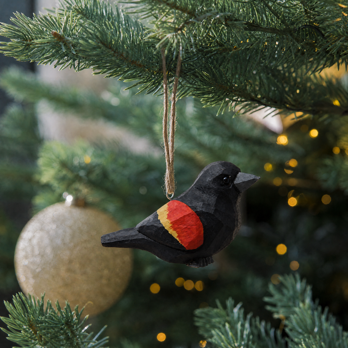 Red-Winged Blackbird wooden ornament hanging by natural twine from a Christmas tree branch, with warm string lights and a gold glitter ball ornament behind it.