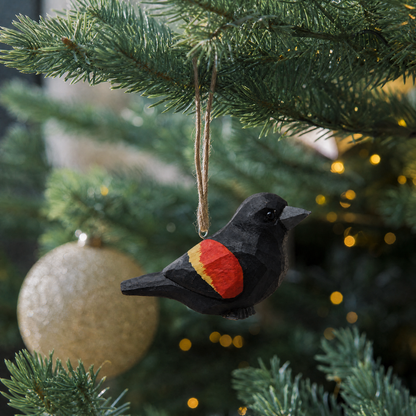 Red-Winged Blackbird wooden ornament hanging by natural twine from a Christmas tree branch, with warm string lights and a gold glitter ball ornament behind it.