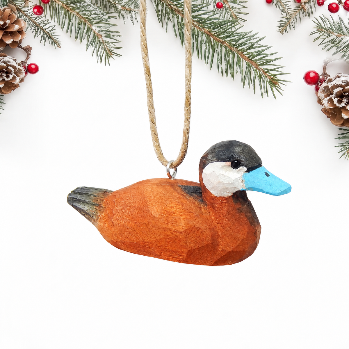 Hand-carved wooden Ruddy Duck ornament with an orange brown body, black cap, white cheek and a sky blue bill, hanging by twine from a silver eye hook in the middle of its back, on a white background with pine branches.