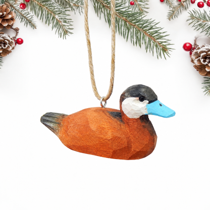 Hand-carved wooden Ruddy Duck ornament with an orange brown body, black cap, white cheek and a sky blue bill, hanging by twine from a silver eye hook in the middle of its back, on a white background with pine branches.