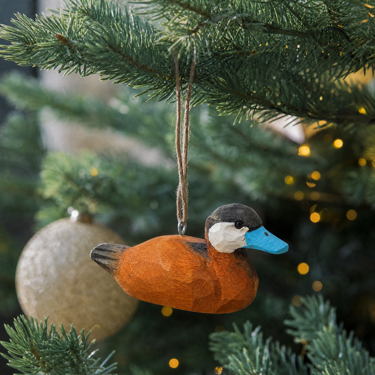 Ruddy Duck wooden ornament hanging by natural twine from a Christmas tree branch, with warm string lights and a gold glitter ball ornament behind it.