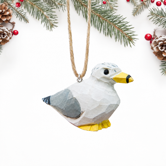 Hand-carved wooden Seagull ornament with a white body, gray wings, a speckled head and a yellow beak and feet, hanging by twine from a silver eye hook in the middle of its back, on a white background with pine branches.